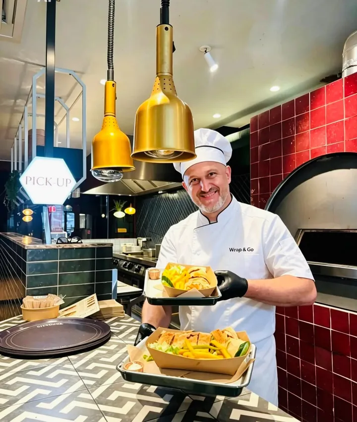 Samy, chef Wrap & Go, présentant deux plateaux dans la cuisine ouverte face aux lampes chauffantes dorées